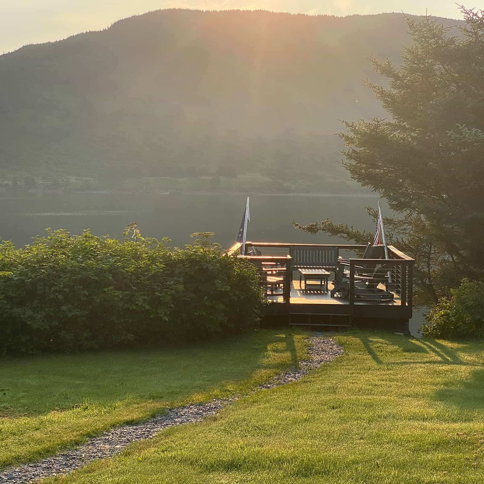 Scenic view of a lakeside deck with chairs and a table at sunset, surrounded by trees and mountains.